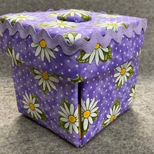 May include: A square fabric box with a lid, decorated with a daisy pattern on a purple polka dot background. The lid has a scalloped edge and a daisy-shaped button. The box is likely used for storage or decoration.