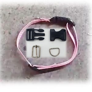May include: A light pink nylon strap collar featuring a black plastic side-release buckle, a silver-toned metal rectangular slider, and a silver-toned metal D-ring. The collar is displayed with the buckle and hardware components detached.