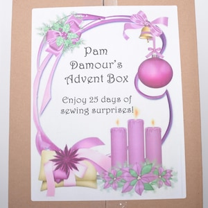 May include: A rectangular cardboard box featuring a white label with the text Pam Damour's Advent Box Enjoy 25 days of sewing surprises! The label includes illustrations of pink candles, a pink ornament, a bell, and purple poinsettia flowers.