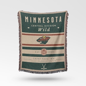 Puede incluir: Una manta rectangular con el texto "MINNESOTA" y "Wild" en un estilo vintage. La manta presenta un logotipo del equipo, rayas y flecos. Los colores son verde, beige y marrón.