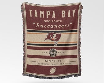 May include: A maroon and tan woven throw blanket featuring the text "TAMPA BAY Buccaneers" and a team logo. The blanket includes the text "EST. 1976" and "RAYMOND JAMES STADIUM". The blanket has a fringed edge.