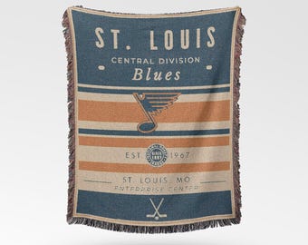 May include: A woven throw blanket featuring the text "ST. LOUIS Blues" in navy blue and tan. The blanket has horizontal stripes in tan and orange, with a logo and text indicating the team's founding in 1967. The blanket has a fringed edge.