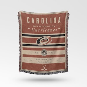 Könnte beinhalten: Eine rechteckige Decke mit dem Text "CAROLINA" oben und "Hurricanes" darunter. Die Decke hat einen rotbraunen Hintergrund mit horizontalen Streifen in Schwarz, Beige und Rotbraun. Das Teamlogo befindet sich in der Mitte.