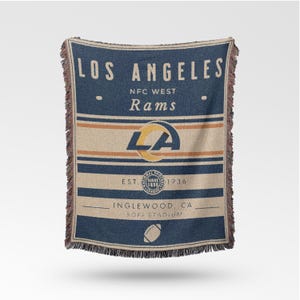May include: A woven throw blanket featuring the text "LOS ANGELES" and "Rams" in navy blue and tan. The blanket includes the team's logo and the text "EST. 1936". The bottom of the blanket reads "INGLEWOOD, CA SOFI STADIUM".