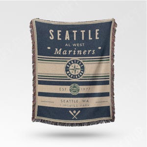 May include: A navy blue and beige throw blanket with the text "SEATTLE Mariners" and a compass logo. The blanket also includes the text "EST. 1977" and "SEATTLE, WA T-MOBILE PARK". The blanket has a fringed edge.