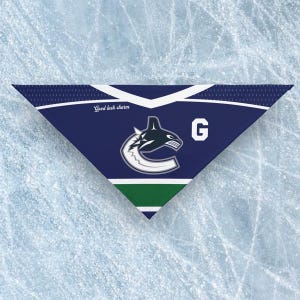 May include: A blue, white, and green triangular bandana featuring the Vancouver Canucks logo and the letter "G". The text "Good luck charm" is printed on the bandana. The bandana is likely made of fabric.