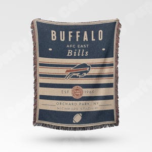 May include: A blue and tan woven throw blanket featuring the text "BUFFALO" and "BILLS". The blanket includes the team's logo and the text "AFC EAST". Additional text includes "EST. SINCE 1960", "ORCHARD PARK, NY", and "HIGHMARK STADIUM".