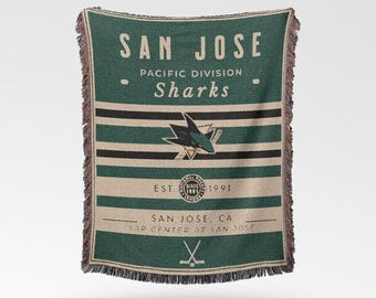 Pode incluir: Cobertor tecido verde e bege com o texto "SAN JOSE Sharks" e um logotipo de tubarão. O cobertor tem listras horizontais e bordas com franjas. O texto "EST. 1991" também é visível.