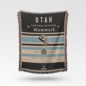 Könnte beinhalten: Eine gewebte Decke in Schwarz, Blau und Beige mit dem Text "UTAH" und "Mammoth". Die Decke hat horizontale Streifen und einen Fransenrand. Das Mammoth-Logo ist deutlich sichtbar.