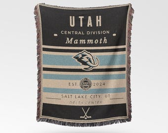 May include: A black and beige woven throw blanket with the text "UTAH" and "Mammoth". The blanket has horizontal stripes in blue and beige, with a fringed edge. The Mammoth logo is visible.