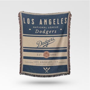 Op de afbeelding: Een geweven deken met het logo van de Los Angeles Dodgers. De deken toont de teamnaam, stadiondetails en de tekst "LOS ANGELES", "Dodgers" en "DODGER STADIUM". Het ontwerp is voornamelijk marineblauw en beige met een vintage logo.