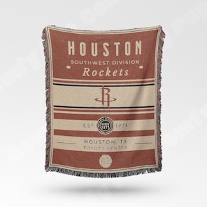 May include: A woven throw blanket featuring the Houston Rockets logo and team name in a vintage style. The blanket is primarily brown and beige, with the team's logo and text prominently displayed. The fringe edges add a classic touch.
