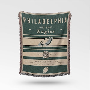 Può includere: Una coperta tessuta verde e beige con la scritta "PHILADELPHIA" e "Eagles". La coperta include il logo della squadra, "NFC East" e "EST. 1933". La parte inferiore recita "PHILADELPHIA, PA, LINCOLN FINANCIAL FIELD".