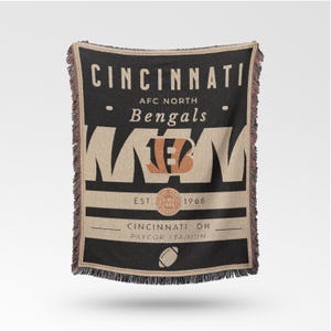 Pode incluir: Uma manta tecida preta e bege com o texto "CINCINNATI" e "Bengals" e o logotipo da equipa. A manta inclui o texto "EST. SINCE 1968" e "PAYCOR STADIUM". As bordas têm uma borda com franjas.