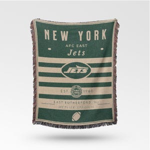 Puede incluir: Manta tejida verde y beige con las palabras "NEW YORK" y "Jets" en blanco. La manta tiene rayas horizontales y un gráfico de fútbol. También es visible el texto "EST. SINCE 1960".