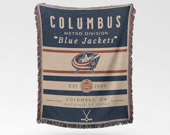 May include: A woven throw blanket featuring the text "COLUMBUS Blue Jackets" in navy blue and tan. The blanket has a fringed edge and includes the team logo and the text "EST. 2000".