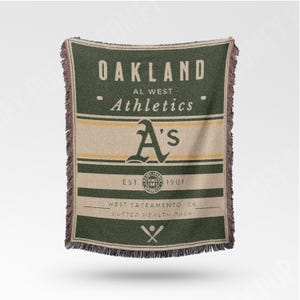 Può includere: Una coperta tessuta verde e beige con la scritta "OAKLAND Athletics" e il logo "A's" della squadra. La coperta ha un bordo con frange e include il testo "EST. 1901" e "WEST SACRAMENTO, CA SUTTER HEALTH PARK".