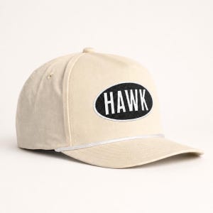 May include: A cream-coloured baseball cap featuring a black oval patch on the front panel. The patch contains the word HAWK in bold white capital letters. A thin white cord runs horizontally across the base of the crown above the brim.