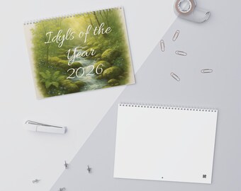 May include: A spiral-bound calendar titled "Idyls of the Year 2026" with a forest stream scene on the cover. The image shows the open calendar with the cover art, and a blank calendar. Office supplies, including paperclips and tape, are scattered around.