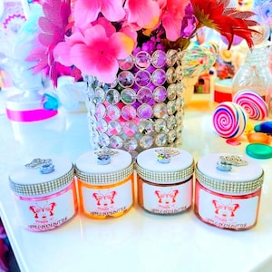 Op de afbeelding: Vier potten Queen C Whipped Body Butter met vlinderlogo's en strass accenten. De potten hebben witte deksels met zilveren kronen. Een vaas met kristallen versieringen bevat roze bloemen. Kleurrijke snoepjes en decoraties op de achtergrond.