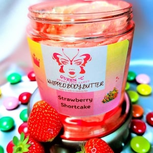 May include: A jar of strawberry shortcake whipped body butter with a pink and yellow label featuring a butterfly design. Fresh strawberries and colourful sweets surround the jar. The jar is on a silver stand.