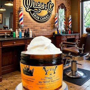 May include: A round amber jar filled with white cream, featuring an orange label with a black crown icon, a butterfly graphic, and the text Queen C My KING Beard Butin. The jar sits on a circular white base in a barbershop setting.