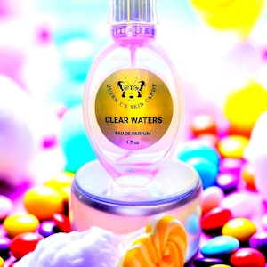 Puede incluir: Un frasco de perfume transparente con una etiqueta dorada que dice "Queen C's Skin Candy Clear Waters Eau de Parfum 1.7 oz." El frasco está sobre un pequeño soporte, rodeado de caramelos coloridos y un caramelo amarillo y blanco.