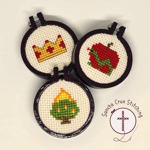May include: Three small embroidery hoops with cross-stitch designs. The hoops are dark brown wood with a white fabric background. The designs are a yellow crown, a red apple with a green snake, and a green tree with orange flames.