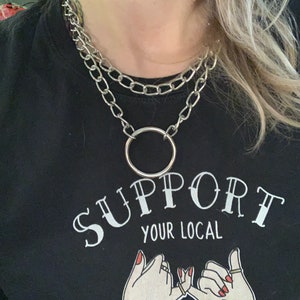 May include: A silver chain necklace with a large silver ring pendant. The necklace is layered and has a chunky chain style. The necklace is worn by a person wearing a black t-shirt with the text "Support Your Local" printed on it.