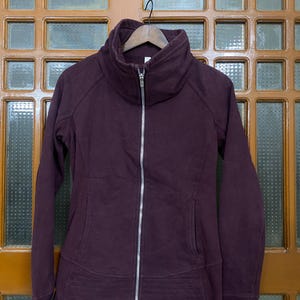 May include: A deep burgundy zip-up jacket with a high collar and ribbed detailing at the waist. The jacket has a full-length silver zipper and two front pockets. The sleeves are long, and the jacket is hanging on a wooden hanger.