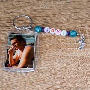 Pode incluir: Um chaveiro de acrílico transparente com uma foto de um homem em uma camiseta branca. O chaveiro tem um chaveiro prateado e um detalhe de contas com o nome "JAVI" escrito em contas de letras brancas. Um pequeno pingente de coala prateado está anexado.