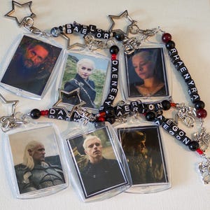 May include: A collection of six rectangular acrylic keychains featuring character portraits from House of the Dragon. Each keychain is attached to a star-shaped clasp and connected by beaded strands with black letter cubes spelling Baelor, Daenerys, Rhaenyra, Daemon, Aemond, and Aegon, accented with red and black beads and silver dragon charms.