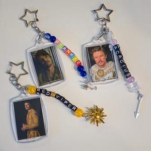 May include: Three rectangular acrylic keychains featuring portraits of a man. Each keychain has a star-shaped ring and a beaded strand. One strand spells FRANKIE with blue beads, one spells ACACIUS with purple beads, and one spells RED VIPER with yellow beads.