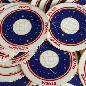 May include: Circular stickers with a space-themed design. Each sticker features a white planet with a grid pattern, surrounded by a blue starry background. The text "STORM" and "GRIMM" are printed in red and white.