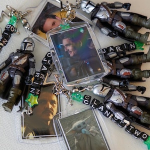 May include: A collection of keychains featuring miniature Mandalorian action figures in silver and brown armour, rectangular photo charms of a man and a small green creature, and black letter beads spelling DJARIN and CLAN OF TWO with green stars.