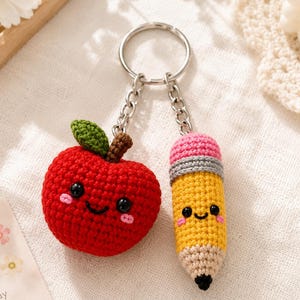 May include: A crochet keychain featuring a red apple with a green leaf and a yellow pencil with a pink eraser, both with embroidered black eyes, smiling mouths, and pink cheeks, attached to a silver metal split ring by two short silver chains.