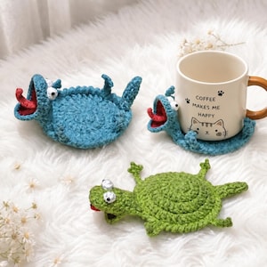 Könnte beinhalten: Gehäkelte Untersetzer in Monsterform in Blau und Grün mit offenen Mäulern und roten Zungen. Einer der Untersetzer hält eine Tasse mit dem Text "Coffee Makes Me Happy" und einer Katzenillustration. Die Untersetzer liegen auf einer weißen, flauschigen Oberfläche.