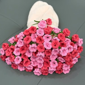 May include: A handmade crochet floral arrangement in shades of pink and red. The flowers are arranged around a folded white towel, creating a decorative display. The crochet work is detailed, showcasing the texture and craftsmanship.