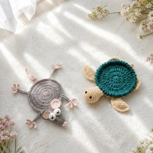 May include: Two handmade crochet coasters shaped like animals. One is a gray mouse with pink inner ears and eyes marked with black crosses. The other is a turtle with a dark teal circular shell and a light beige head and flippers.