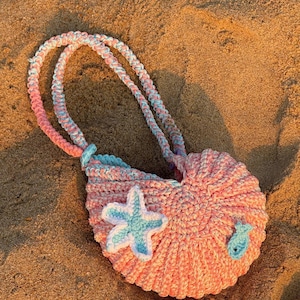 May include: A handmade crochet bag in a seashell shape, resting on sand. The bag is primarily pink with blue and white accents, featuring a blue starfish and fish applique. The handles are a twisted cord in matching colors.
