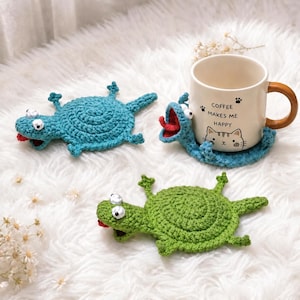 May include: Two crocheted turtle coasters in teal and green, each with googly eyes and a red tongue. A third matching teal turtle coaster is wrapped around a cream-colored ceramic mug featuring a cat illustration and the text COFFEE MAKES ME HAPPY.