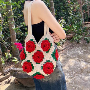 May include: A cream-colored crochet tote bag adorned with vibrant red rose appliqués and green leaves. The bag features a square pattern design and long straps for carrying. The roses are a prominent feature, adding a touch of floral elegance to the handmade accessory.