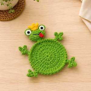 May include: A round green crochet coaster shaped like a frog. The frog features a yellow crown on its head, white and black eyes, and a small red tongue sticking out. It has four small webbed feet extending from the circular green body.