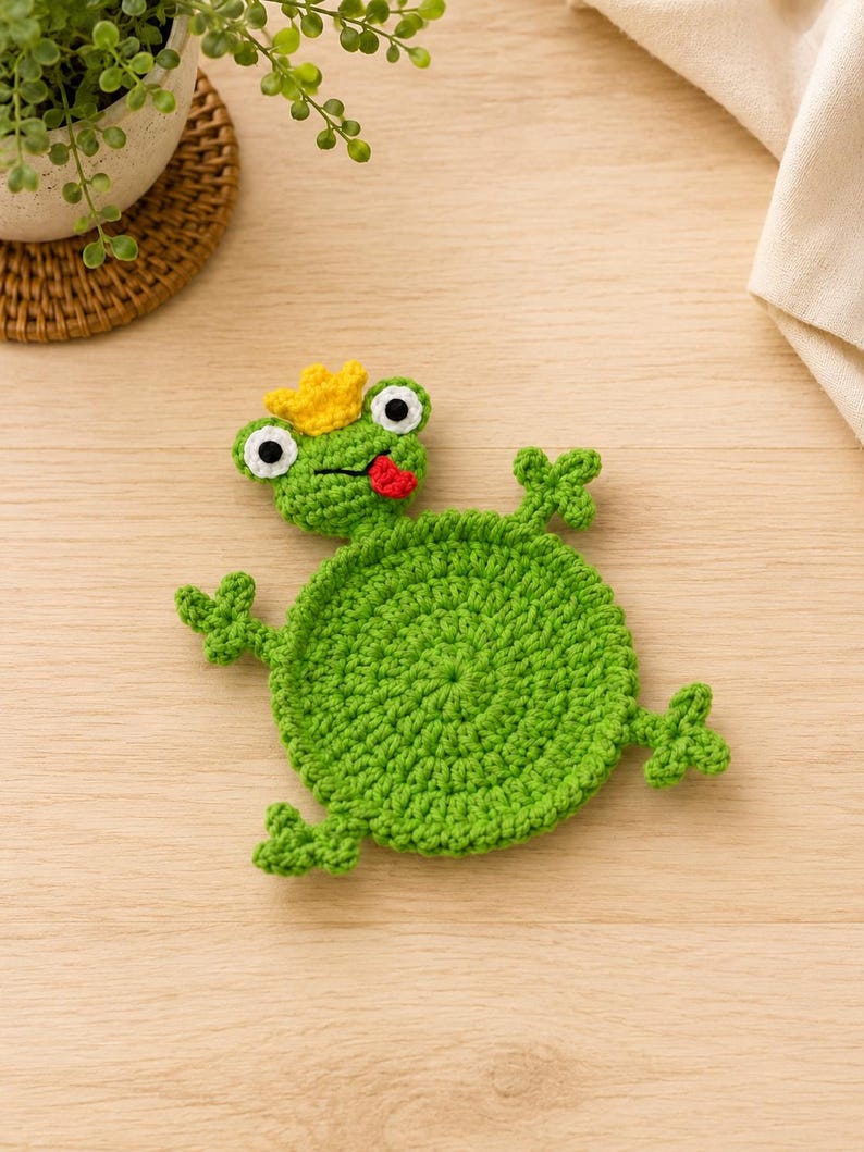 May include: A round green crochet coaster shaped like a frog. The frog features a yellow crown on its head, white and black eyes, and a small red tongue sticking out. It has four small webbed feet extending from the circular green body.