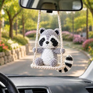 May include: A crocheted raccoon ornament hanging from a rearview mirror. The raccoon has gray and white yarn, black eye patches, and a striped tail. It sits on a small, cream-colored crocheted swing suspended by two thick, twisted cream-colored ropes.