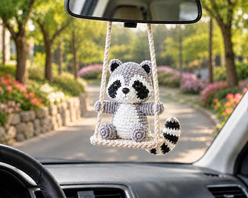 May include: A crocheted raccoon ornament hanging from a rearview mirror. The raccoon has gray and white yarn, black eye patches, and a striped tail. It sits on a small, cream-colored crocheted swing suspended by two thick, twisted cream-colored ropes.