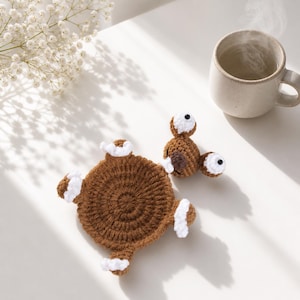 May include: A brown crocheted coaster shaped like a bear with a circular body, four white-tipped paws, and a head featuring two round ears, two white eyes with black pupils, and a dark brown nose, positioned near a steaming ceramic mug.
