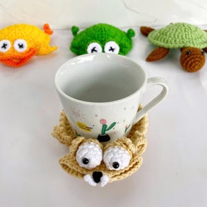 May include: A white ceramic mug with a small cactus illustration and the text "STAY AWAY FROM ME" sits on a beige crocheted coaster shaped like a creature with large white eyes. In the background are three crocheted toys: a yellow duck, a green frog, and a green turtle.
