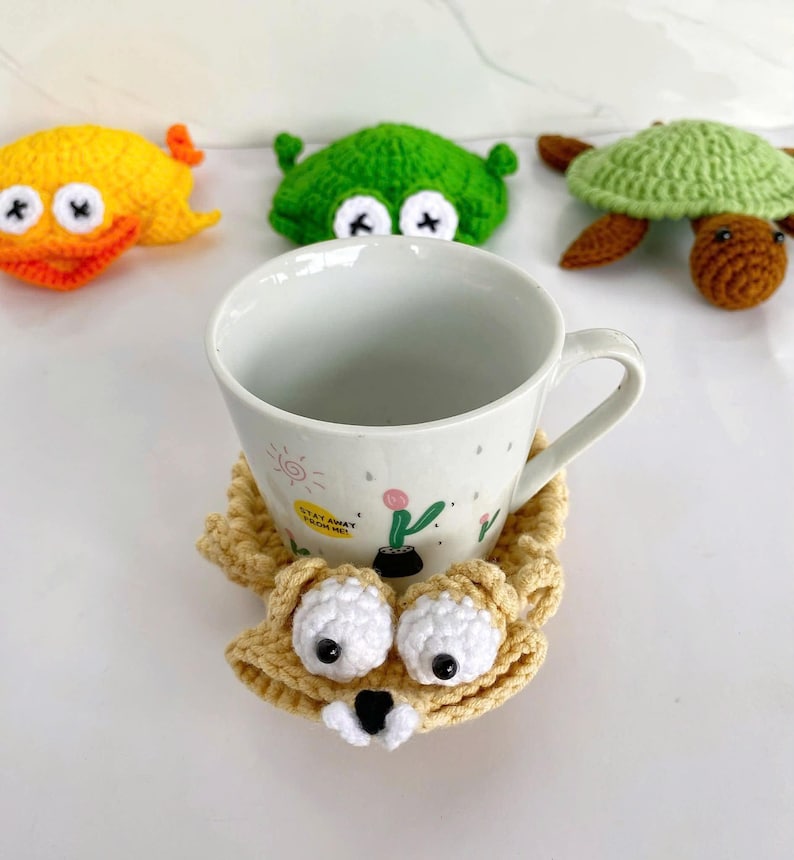 May include: A white ceramic mug with a small cactus illustration and the text "STAY AWAY FROM ME" sits on a beige crocheted coaster shaped like a creature with large white eyes. In the background are three crocheted toys: a yellow duck, a green frog, and a green turtle.