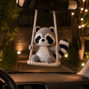 May include: A crocheted raccoon ornament hanging from a rearview mirror. The raccoon is grey, white, and black with a striped tail, sitting on a small, beige crocheted swing suspended by two thick, twisted ropes attached to the mirror mount.